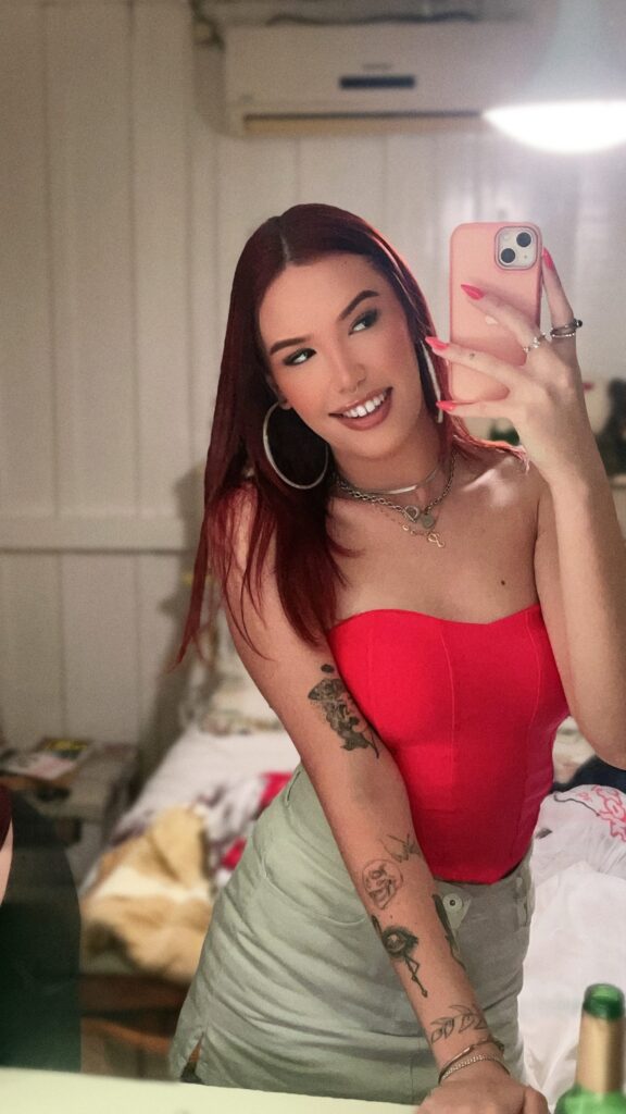 Young woman with red hair taking a mirror selfie in a bedroom, wearing a red strapless top, light green shorts, hoop earrings, and layered necklaces, with visible arm tattoos.